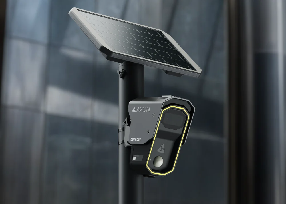 Axon's Outpost camera mounted on a black pole under a solar panel, with the Axon and Outpost names on its gray housing and a yellow light around its lens.